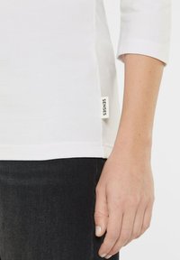 Weißes T-Shirt mit drei Viertelärmeln aus weicher Baumwolle, mit einem Seitenlabel "SENSES." Kombiniert mit dunklen Jeans.