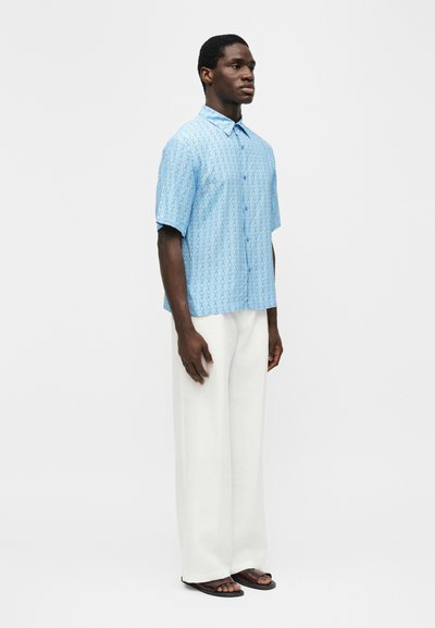 Άνδρας που φοράει ανοιχτό μπλε πουκάμισο με κοντό μανίκι και σχέδια, άσπρο παντελόνι φαρδύ στα πόδια και μαύρα σανδάλια, στέκεται μπροστά σε ένα απλό φόντο.