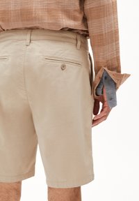 Shorts beiges à la texture lisse, avec deux poches arrière et un détail de bouton unique. Portés avec une chemise à carreaux brune à boutons.