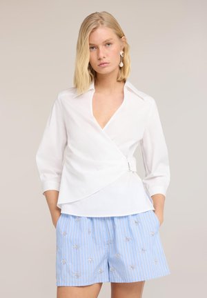 Jonge vrouw met kort blond haar, die een witte wikkelblouse en lichtblauwe gestreepte shorts met kleine bloembroderijen draagt, staat met handen in de zakken.