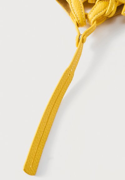 Κίτρινο λουρί από δέρμα τσάντας με ορατές ραφές και μεταλλικό πριτσίνι, μερικώς τυλιγμένο σε λευκό φόντο.