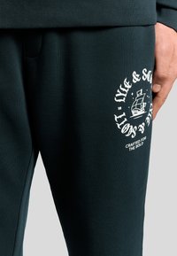 Donkergroene sweatpants van zacht materiaal, met een witte grafiek van een schip en de tekst "Live & Sport" op de dij. Geribbelde tailleband.