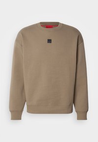 Felpa beige con collo rotondo, maniche lunghe, polsini e orlo a costine, e una piccola patch nera al centro del petto.