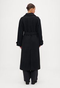 Garšs melns mētelis, izgatavots no teksturēta auduma, ar plašu apkakli, jostu viduklī un pogām pie aprocēm, taisnas siluets un vidussakaru garums.