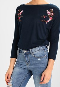 Marineblauw long-sleeve shirt met geborduurde roze bloemenpatronen op de borst, gecombineerd met lichtblauwe distressed skinny jeans.