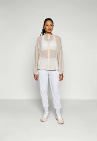 Lätt, genomskinlig beige jacka med hög krage och framdragkedja, kombinerad med vita sweatpants och beigea träningsskor.