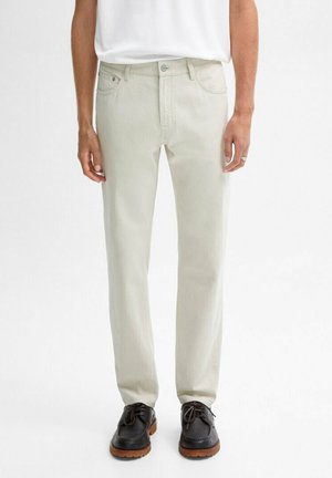 Homme portant un jean droit écru, une chemise blanche rentrée et des chaussures en cuir noir avec semelles marron, debout devant un fond uni.