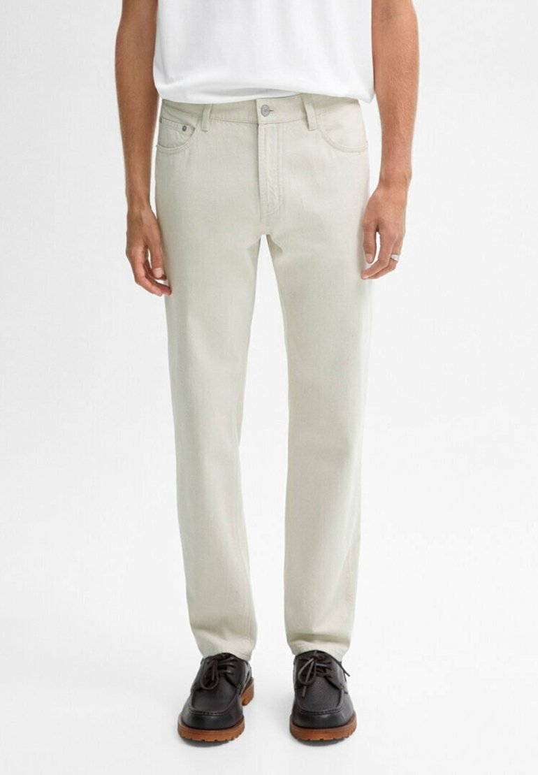 Homme portant un jean droit écru, une chemise blanche rentrée et des chaussures en cuir noir avec semelles marron, debout devant un fond uni.