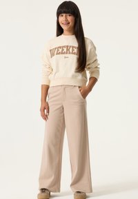 Sudadera crema con estampado "WEEKEND", combinada con pantalones de pierna ancha de rayas finas en beige claro. La modelo está de pie con la mano en el bolsillo, luciendo zapatillas deportivas.