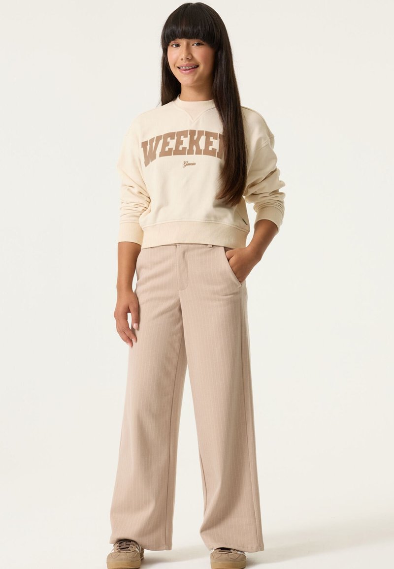 Sudadera crema con estampado "WEEKEND", combinada con pantalones de pierna ancha de rayas finas en beige claro. La modelo está de pie con la mano en el bolsillo, luciendo zapatillas deportivas.