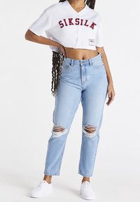 Frau in einem weißen, kurzärmeligen, knöpfbaren Hemd mit "SIKSILK"-Aufschrift, hellblauen, zerrissenen Jeans und weißen Sneakers, die mit einer Hand auf der Hüfte steht.