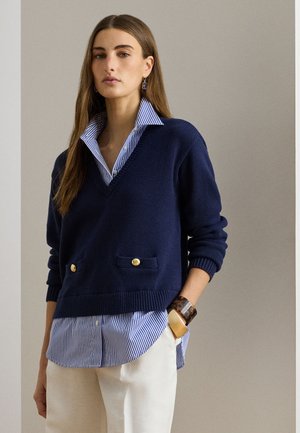 Γυναίκα που φοράει πουλόβερ μπλε ναυτικό με V-λαιμόκοψη πάνω από μπλε ριγέ πουκάμισο με κουμπιά, λευκό παντελόνι και χοντρό βραχιόλι σε σχέδιο χελώνας και χρυσό.