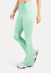 Leggings évasés en tissu vert menthe, texture lisse, taille haute, avec coutures latérales, assortis à des chaussures de sport blanches.
