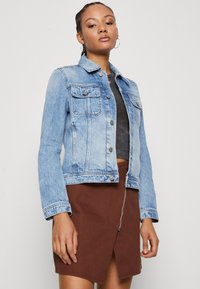 Veste en denim bleu clair avec une coupe classique, deux poches poitrine et des boutons en métal argenté. Associée à une jupe zippée marron.