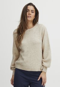 Beige neulepaita pyöreällä kaula-aukolla, puffattuja pitkiä hihoja, koristeltu pienillä valkoisilla helmillä, ribbatut hihansuut ja pehmeä rakenne.