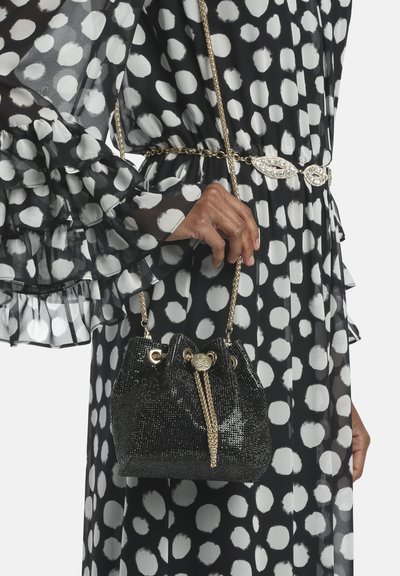 Sort paliet-bøtte taske med guld kæde strop; har snørelukning og metalliske detaljer. Bæres med en sort og hvid prikket kjole.