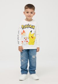 T-shirt bianco a maniche lunghe con grafiche colorate di Pokémon, abbinato a jeans strappati blu chiaro e sneakers bianche.