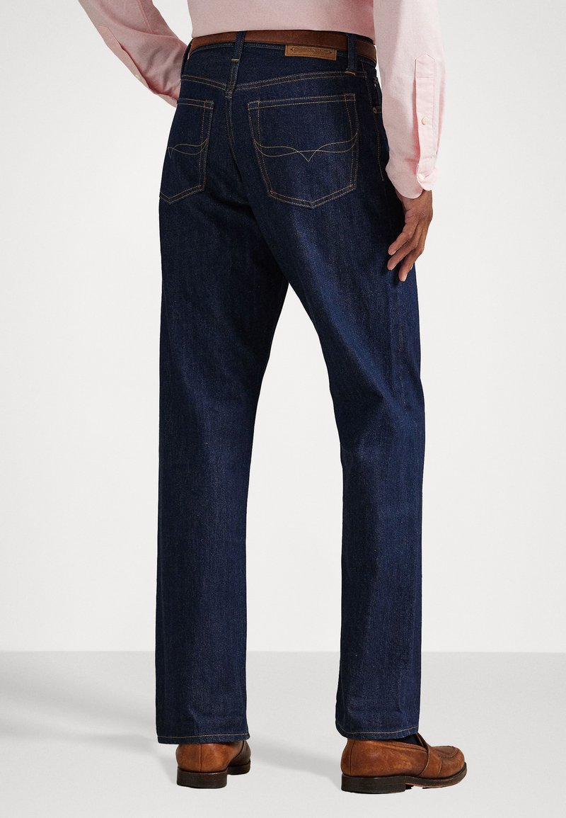 Dark blue denim jeans with straight legs, visible rear pockets, and subtle stitching details. Worn with brown shoes and a light shirt.
