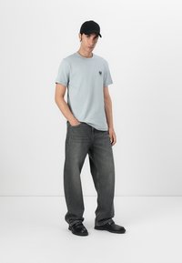 T-shirt de algodão azul claro com decote redondo e pequeno logótipo, combinada com jeans de perna larga cinza escuro e sapatos pretos. Um boné preto acrescenta detalhe.