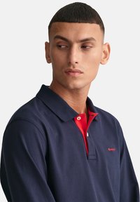 Tengerészkék pólóing piros gallérral. Az anyag puhanak tűnik, sima textúrával. Kis logó található a mellkason, kontrasztos gombokkal.