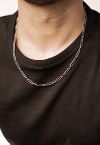 Silver metal necklace featuring elongated links with intersections, worn over a black ribbed shirt. Textured surface and visible sheen.