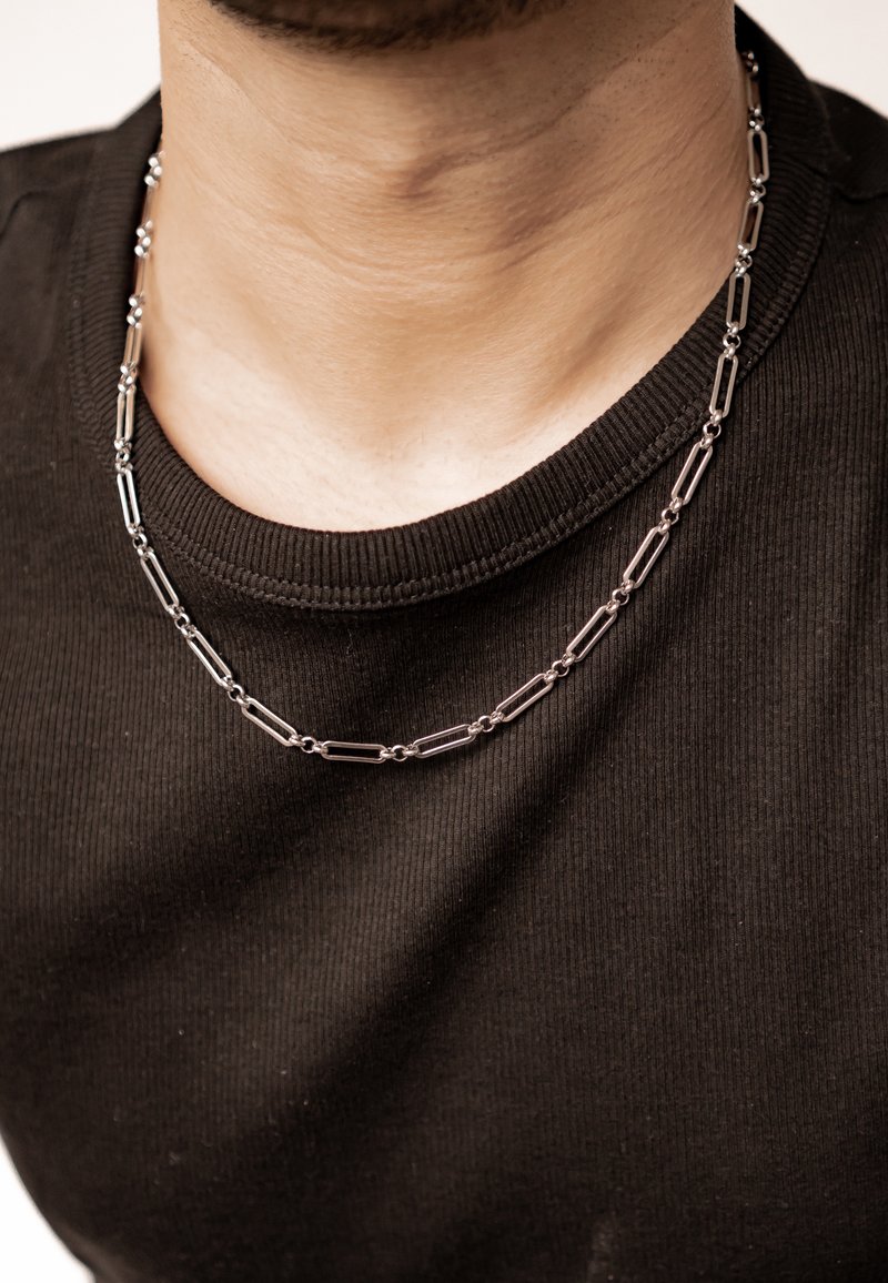 Silver metal necklace featuring elongated links with intersections, worn over a black ribbed shirt. Textured surface and visible sheen.