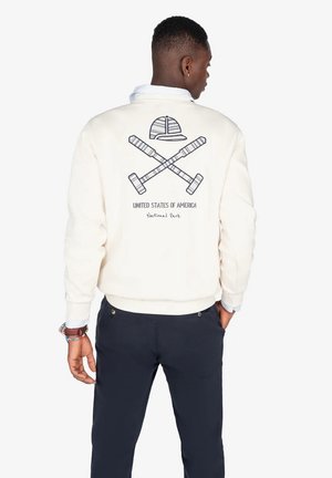 Sudadera color crema con un diseño en la parte posterior que presenta murciélagos cruzados y un sombrero, etiquetada como "Estados Unidos de América" y "Parque Nacional".