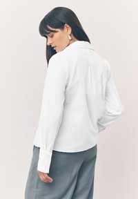 Chemise blanche à manches longues avec une coupe ajustée, détails de poignets à boutons, et une couture au dos. Portée avec un pantalon gris à coupe ample.