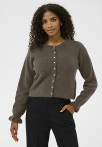 Ženska s kodrastimi lasmi nosi rjav puhast kardigan s bisernimi gumbi in črne hlače, stoji z roko v žepu pred belo ozadje.