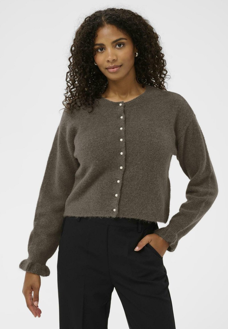 Ženska s kodrastimi lasmi nosi rjav puhast kardigan s bisernimi gumbi in črne hlače, stoji z roko v žepu pred belo ozadje.