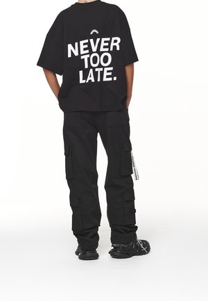 Persona que lleva pantalones cargo negros y una camiseta negra oversized con texto blanco "NEVER TOO LATE" visible en la espalda.