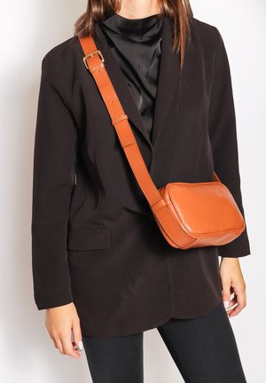 Borsa a tracolla in pelle marrone con forma rettangolare, texture liscia, tracolla regolabile e dettaglio in fibbia metallica, abbinata a un blazer nero.