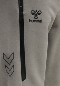 Grijze sportieve broek met een gestructureerde stof, zwarte ritssluiting en Hummel-logo; met zwarte chevron-accenten aan de zijkant.