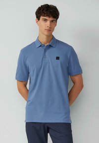 Blaues Poloshirt aus weicher Baumwolle, mit einem Drei-Knopf-Verschluss und einem kleinen schwarzen Logopatch auf der linken Brust. Kurze Ärmel.