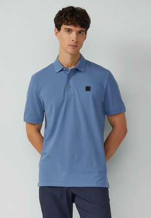 Blaues Poloshirt aus weicher Baumwolle, mit einem Drei-Knopf-Verschluss und einem kleinen schwarzen Logopatch auf der linken Brust. Kurze Ärmel.