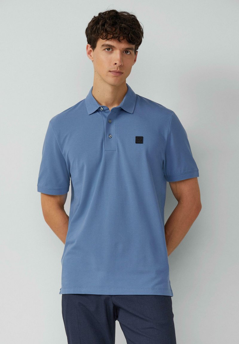 Blaues Poloshirt aus weicher Baumwolle, mit einem Drei-Knopf-Verschluss und einem kleinen schwarzen Logopatch auf der linken Brust. Kurze Ärmel.
