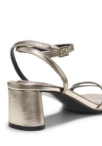 Μεταλλικού χρυσού χρώματος σανδάλι με χοντρό τακούνι, λεπτά λουράκια στον αστράγαλο και τα δάχτυλα, κούμπωμα με χρυσή αγκράφα και μαύρη σόλα.