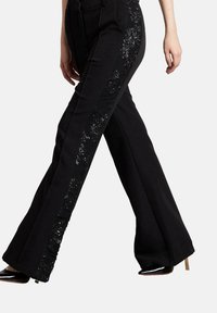 Pantalones negros con forma acampanada, con acentos de lentejuelas en los laterales, tela suave y cintura ajustada. Se ven parcialmente unos zapatos de tacón alto.