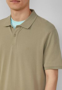 Beige polo shirt van een gestructureerde stof, met een sluiting van twee knopen, korte mouwen en subtiele branding op de borst. Gelaagd over een lichtblauw shirt.