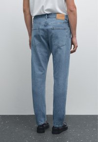 Světlé modré denimy s uvolněným střihem, mají dvě zadní kapsy a malou hnědou koženou nášivku na pasu. Hladká textura.