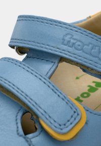 Δερμάτινο σανδάλι ανοιχτού γαλάζιου χρώματος με δύο ρυθμιζόμενους ιμάντες, κίτρινες λεπτομέρειες και σχέδιο με κοψίματα. Το εσωτερικό διαθέτει μπεζ φόδρα και λογότυπο.