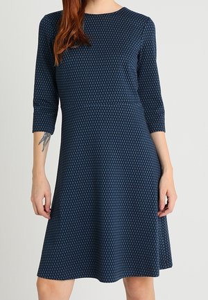 Kvinde iført en knælængde marineblå kjole med et lille diamantmønster og trekvartærmer, stående mod en ensfarvet lys baggrund.