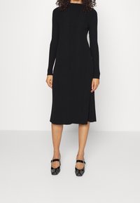 Robe noire en tricot côtelé avec des manches longues et un col montant, présentant une coupe ajustée et une longueur au genou. Associée à des chaussures noires brillantes.
