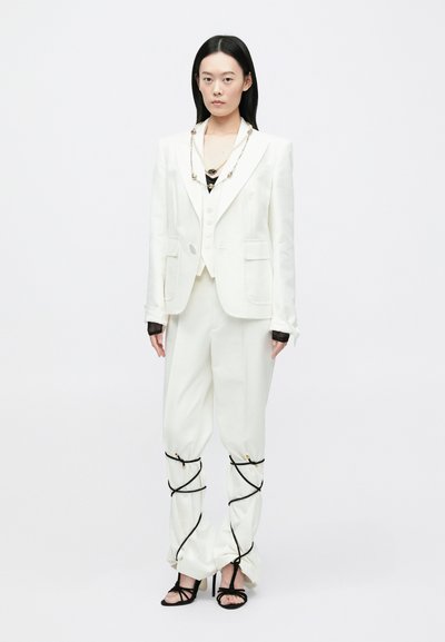 Vrouw in een wit op maat gemaakt pak met geplooide broek gestrikt met zwarte koorden en zwarte high heels met bandjes, staand tegen een witte achtergrond.