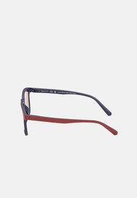 Gafas de sol rectangulares con un marco azul marino y brazos rojos. Fabricadas en un 85% de plástico PET, con un acabado mate y un logotipo elevado.