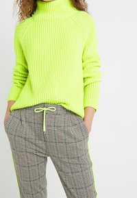 Neonowozielony prążkowany sweter z golfem w zestawieniu z szarymi, w kratę spodniami z neonowozielonymi akcentami i talią na sznurek.