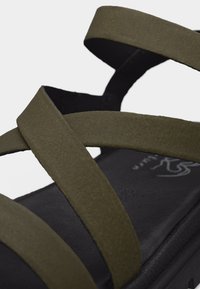 Κοντινό πλάνο μαύρης πλατφόρμας σανδάλια με φαρδιές χακί λουριά που διασταυρώνονται πάνω από το πόδι και γύρω από τον αστράγαλο.