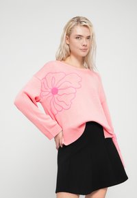 Ροζ πλεκτό πουλόβερ με μεγάλο σχέδιο λουλουδιού σε σκουρότερο ροζ χρώμα, με μακριά μανίκια και στρογγυλή λαιμόκοψη, συνδυασμένο με μαύρη φούστα.