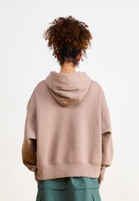 Luźna bluza z kapturem z miękkiego beżowego materiału, z kapturkiem na sznurek i ściągaczem, zestawiona z zieloną warstwową spódnicą, widziana od tyłu.