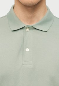 Κοντινό πλάνο ενός ατόμου που φοράει ανοιχτό πράσινο πόλο πουκάμισο με δύο κουμπιά κλειστά στο γιακά και το επάνω μέρος του στήθους.